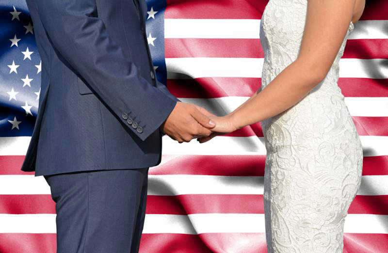 A-bride-and-groom-from-the-shoulders-down, facing each other in front of an American flag.